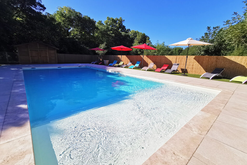 Piscine extérieure aux eaux bleues et blanches sous un ciel bleu, entourée de chaises longues colorées et de parasols rouges et blancs.